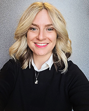 White woman with blonde shoulder length hair wearing a black sweater over a white collar. Silver Jayhawk necklace.